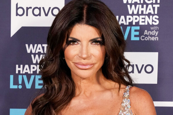 Teresa smiling with full glam wearing a crystal embellished jumpsuit in front of the WWHL step and repeat.