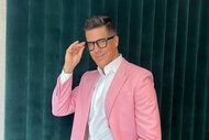 Fredrik Eklund posing in front of a deep green, velvet, backdrop wearing black framed glasses and a pink blazer with a white buttoned down shirt.