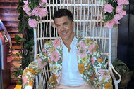 Fredrik in a floral blazer sitting in a white, cane, chair with pink roses.