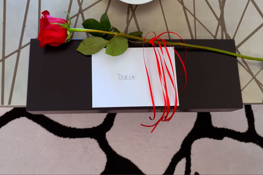 A black box with a white envelope and rose atop it is set on a table