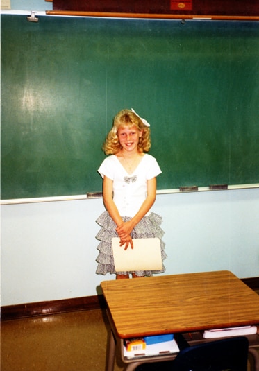 A young Gretchen Rossi standing in front of a black board.