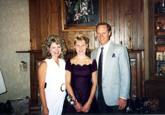 A young Gretchen Rossi standing smiling with her parents.