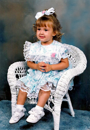 Ashlee Holmes as a child in an Easter dress and bow sitting in a white wicker chair