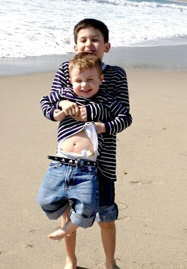 CJ Laurita holding up his brother Nicholas in matching blue striped shirts on the beach as children