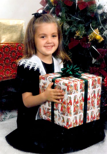 Ashlee Holmes as a child in a Christmas dress holding a present in front of a tree