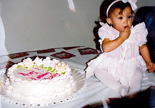 A baby Ashlee Holmes in a white dress and bow next to a large white birthday cake