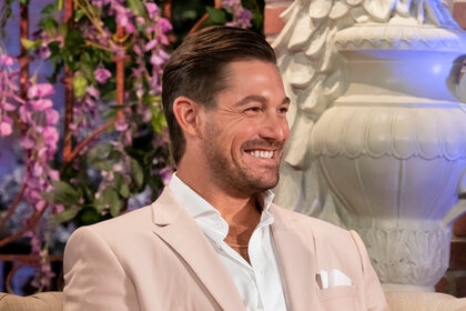 Craig Conover smiling in a pink suit in front of a large, white, statue and florals.