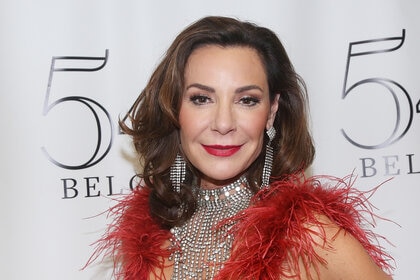 Luann in a red, feathered, outfit in front of a step and repeat.