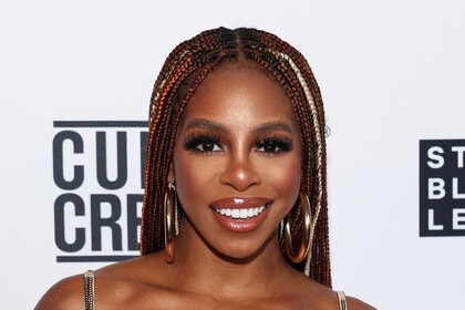 Candiace smiling wearing braids and a sparkly, silver, bralette in front of a step and repeat.