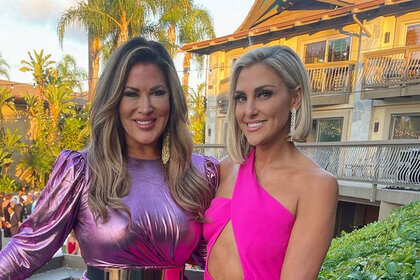 Emily Simpson and Gina Kirschenheiter smile in pink outfits on a balcony.