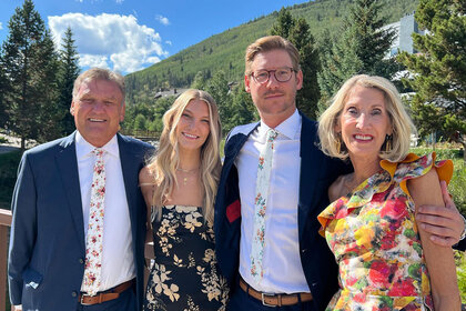 Austen Kroll posing in cocktail attire with his family outdoors in front of a forest with mountains.