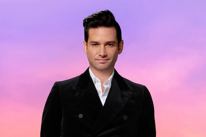 Josh Flagg wearing a black blazer in front of a purple and pink background.
