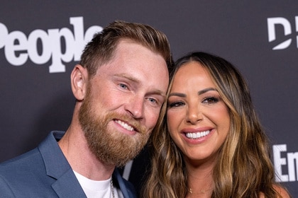 Kristen Doute and Luke Broderick smile together in front of a step and repeat at an event.