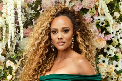 Ashley Darby posing in an emerald green gown in front of a backdrop of flowers.