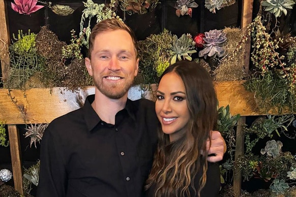 Kristen Doute and Luke Broderick hugging and posing together in front of a plant wall.