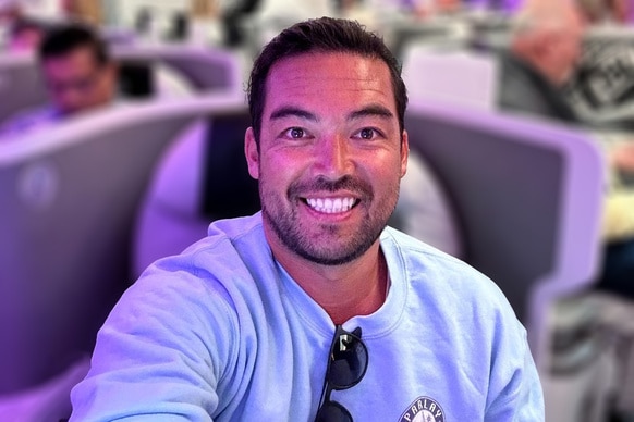 Colin Macrae wearing a blue shirt, smiling while sitting in first class on a flight