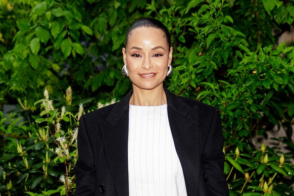 Sai De Silva smiling with her hair slicked back, wearing a white dress and a black blazer in front of shrubs