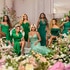 The posing in an emerald green gown in front of a backdrop of flowers.