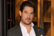 Craig Conover wearing a grey suit standing in front of a rustic seating area.