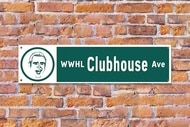 A street sign with a cartoon of Andy Cohen's face advertising the WWHL clubhouse on a brick wall