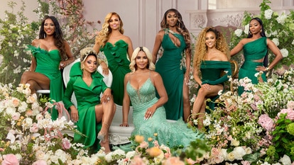 The posing in an emerald green gown in front of a backdrop of flowers.