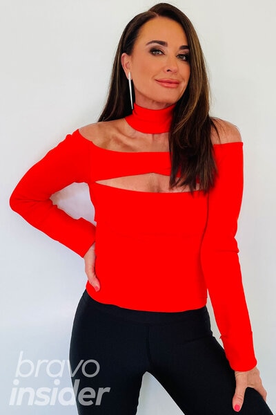 Kyle Richards smiling and posing in a bright red, cutout, top in front of a white wall.