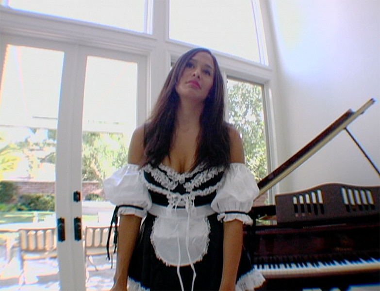 Jo De La Rosa wearing a maid outfit and cleaning a piano.