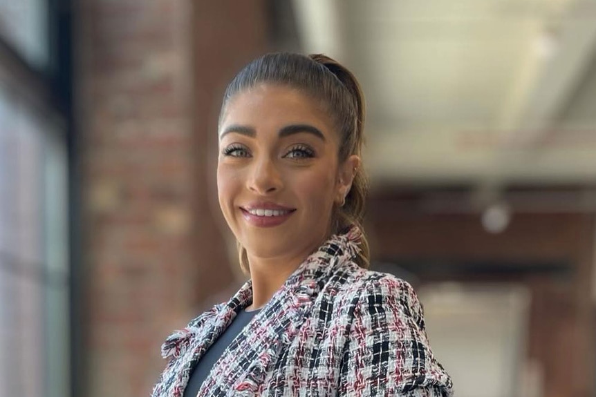 Gia in a tweed blazer standing by windows.