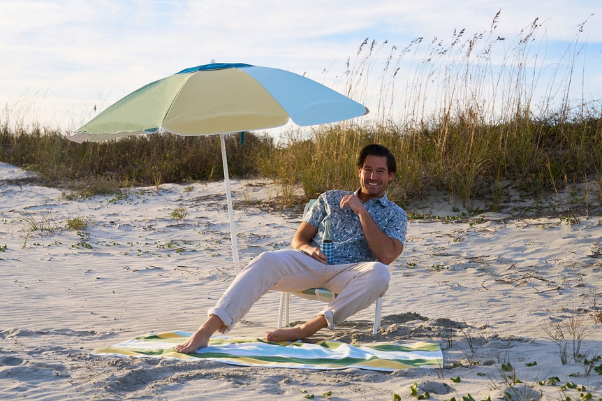 Craig Conover sitting on a beach with an umbrella and blanket.