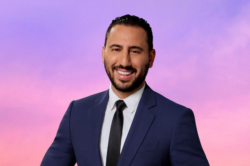 Josh Altman wearing a blue blazer in front of a purple and pink background.
