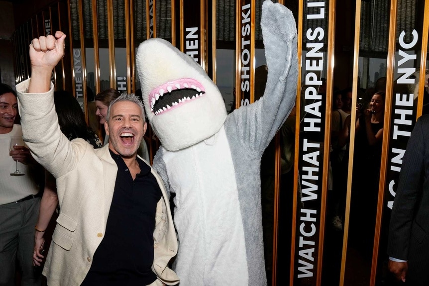 Andy Cohen and a person in a shark costume pumping their fists in the air