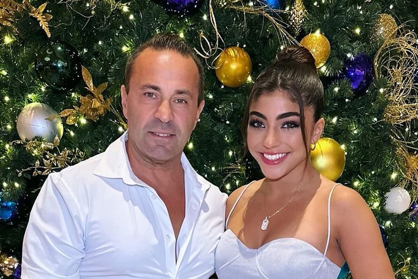 Joe Giudice and Milania Giudice smiling together in front of a Christmas tree.