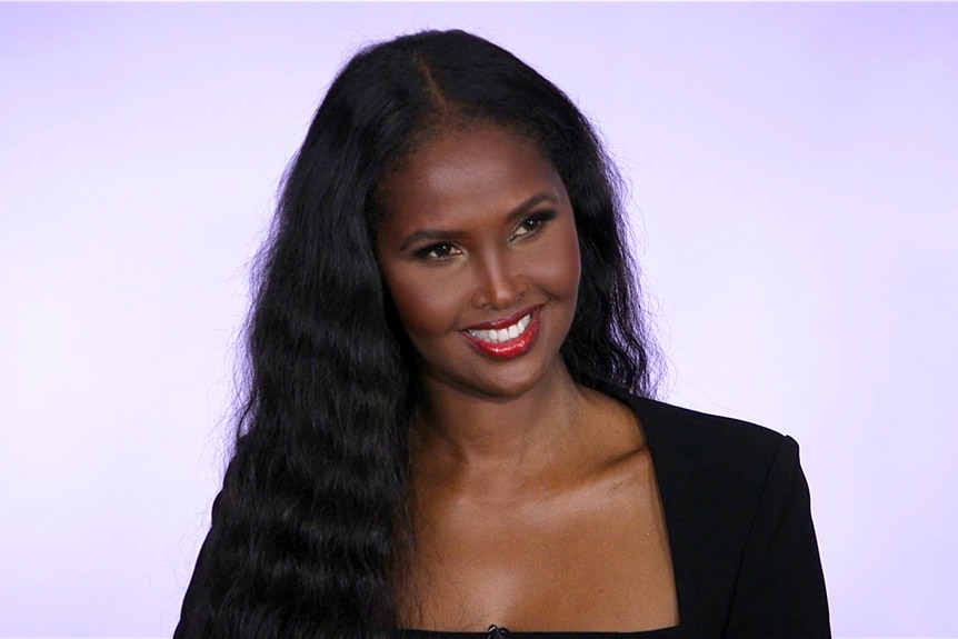 Ubah Hassan wearing a black blazer in front of a purple backdrop.